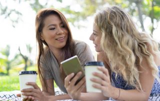 Cheerful young ladies drinking coffee and using gadget together